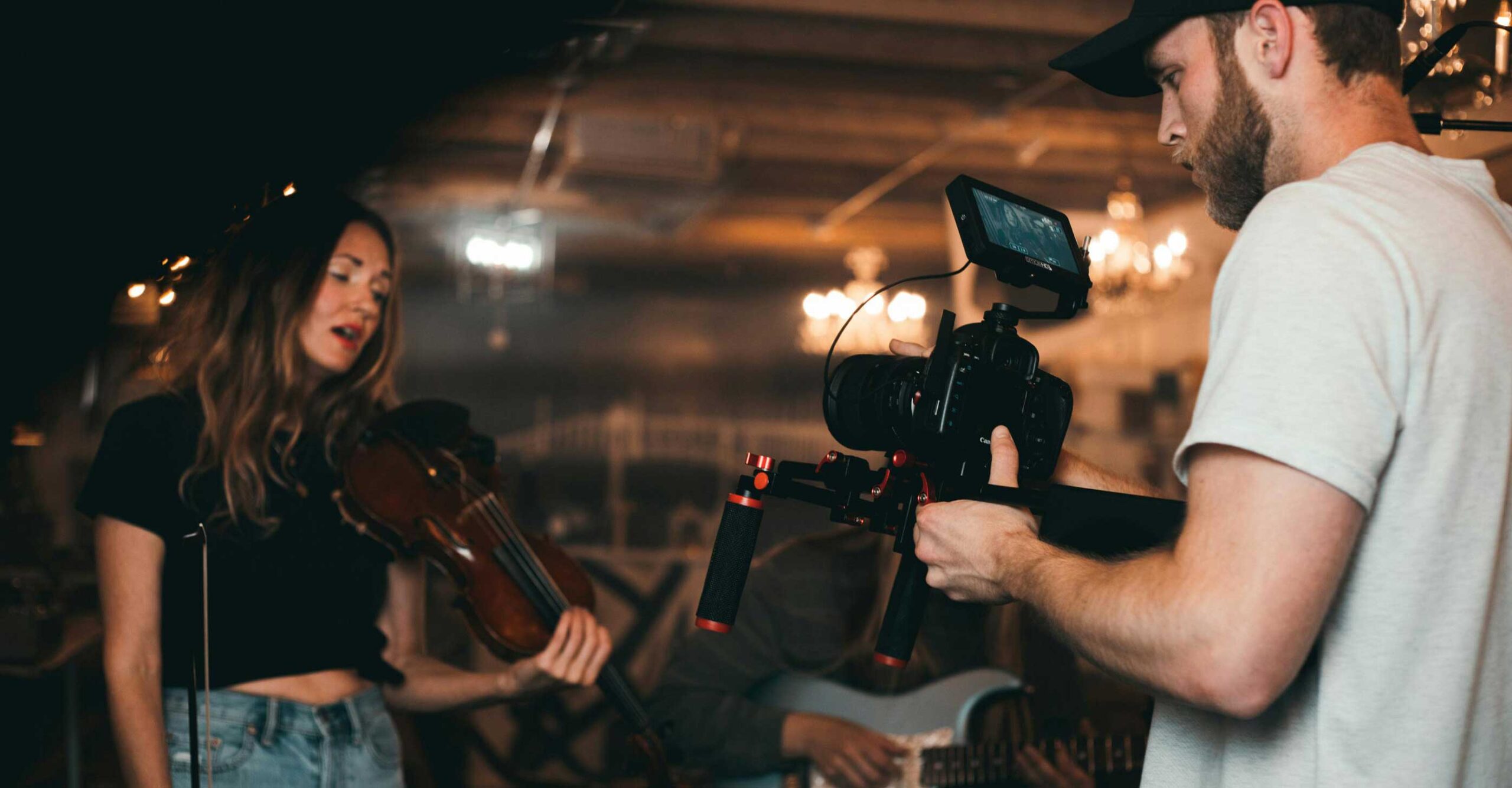 A violinist sings while being filmed by a videographer holding a camera.